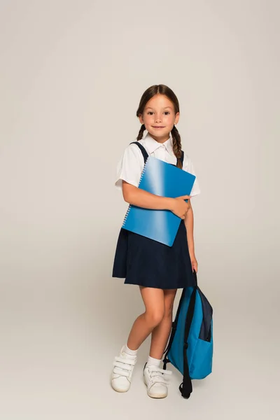 Ganzkörperansicht eines lächelnden Schulmädchens, das mit blauem Rucksack und Kopierbüchern auf grau steht — Stockfoto