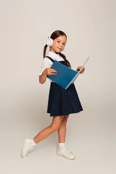 Full length view of schoolgirl in headphones holding copy book on grey — Stock Photo