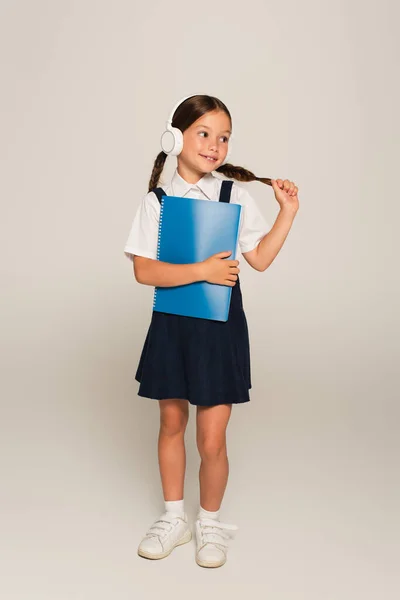 Positive schoolgirl in headphones touching pigtail while holding copy book on grey — Stock Photo