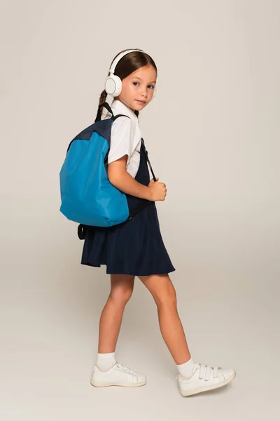 Colegiala en auriculares caminando con mochila azul y mirando a la cámara en gris - foto de stock