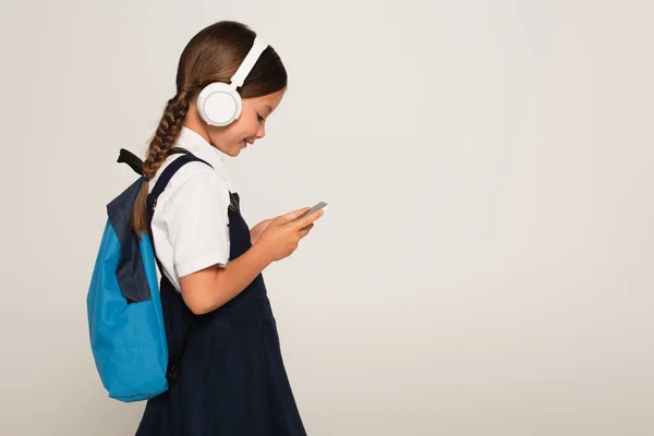 Side view of cheerful schoolkid in headphones using smartphone isolated on grey — Stock Photo