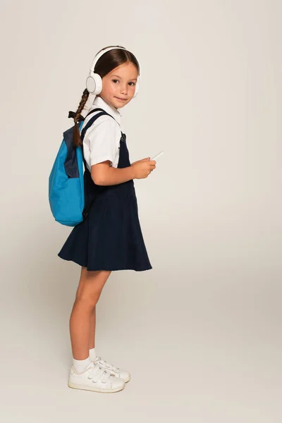 Full length view of schoolgirl in headphones holding mobile phone on grey — Stock Photo