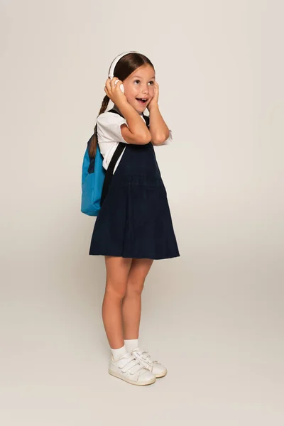 Full length view of excited schoolgirl touching headphones while looking away on grey — Stock Photo