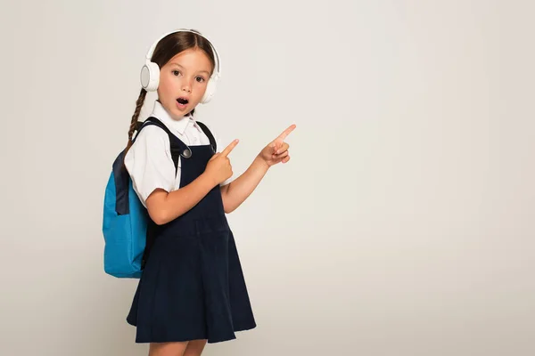 Colegial asombrado en auriculares apuntando con los dedos aislados en gris - foto de stock