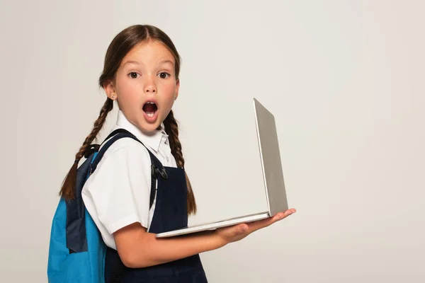 Здивована школярка з відкритим ротом дивиться на камеру, тримаючи ноутбук ізольований на сірому — стокове фото