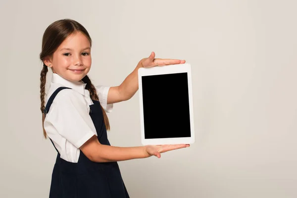 Улыбающийся школьник показывает цифровой планшет с чистым экраном, изолированным на сером — стоковое фото