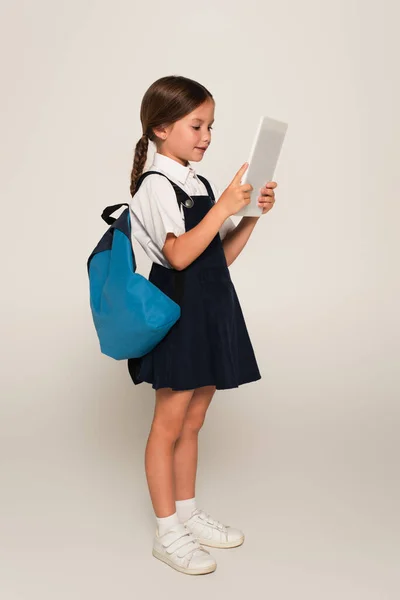 Повний вигляд школярки з синім рюкзаком з використанням цифрового планшета на сірому — стокове фото