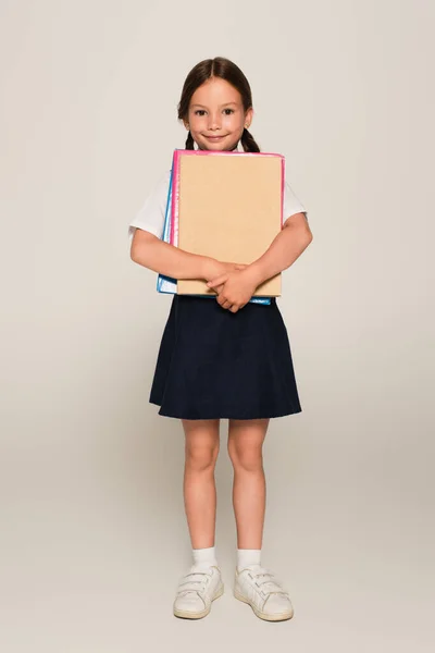 Full length view of schoolgirl with notebooks looking at camera on grey — Stock Photo