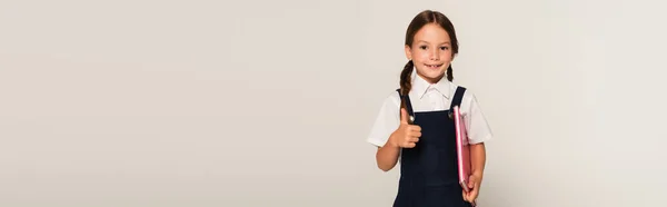 Pleased schoolgirl holding notebook and showing thumb up isolated on grey, banner — Stock Photo