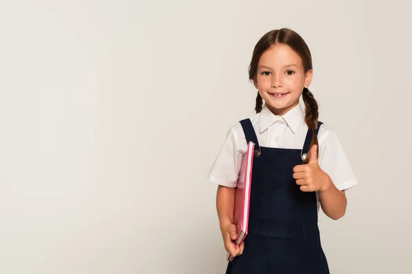 Colegial positivo con libro de copias que muestra el pulgar hacia arriba aislado en gris - foto de stock