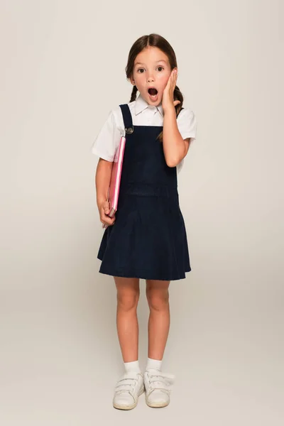 Shocked schoolgirl with open mouth touching face while looking at camera on grey — Stock Photo