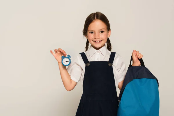 Smiling schoolgirl holding blue backpack and small alarm clock isolated on grey — Stock Photo