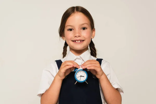 Fröhliches Schulkind mit kleinem Wecker, während es isoliert auf grau schaut — Stockfoto