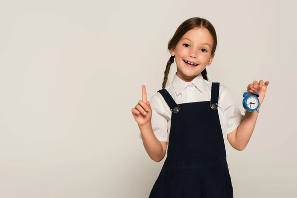 Écolier heureux montrant petit réveil et pointant du doigt isolé sur gris — Photo de stock