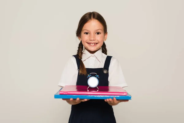 Радісна школярка тримає блокноти з невеликим будильником ізольовані на сірому — стокове фото