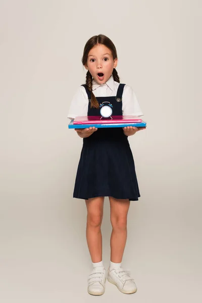 Menina espantada no uniforme da escola segurando cadernos e pequeno despertador no cinza — Fotografia de Stock