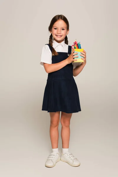 Joyeuse écolière tenant porte-stylo avec crayons et stylos en feutre sur gris — Photo de stock