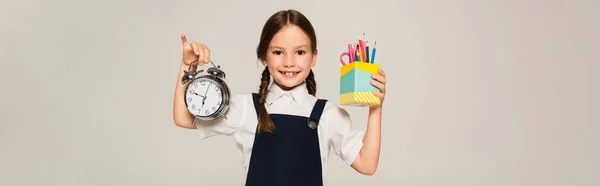 Scolaretta con grande sveglia e portapenne sorridente alla telecamera isolata su grigio, banner — Foto stock