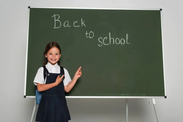 Радісна дівчина в шкільній формі вказуючи на крейдяну дошку зі спиною до школи малювання на сірому — стокове фото