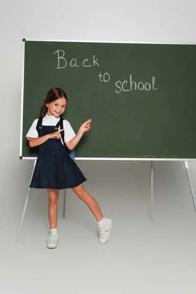 Vista completa de la colegiala apuntando a la pizarra con letras de vuelta a la escuela en gris - foto de stock