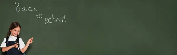 Щаслива школярка вказує на крейдяну дошку зі спиною до шкільного написання, банер — стокове фото