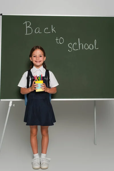 Allievo soddisfatto che tiene il portapenne vicino alla lavagna con iscrizione torna a scuola su grigio — Foto stock