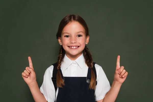 Joyful girl in school uniform pointing with fingers near chalkboard while looking at camera — Stock Photo