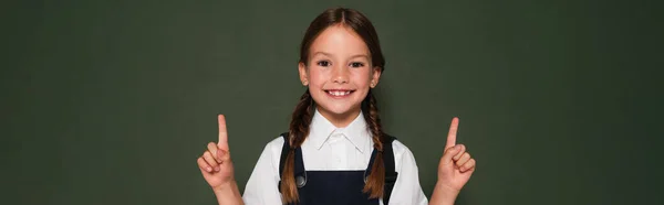 Cheerful schoolgirl looking at camera while pointing with fingers near chalkboard, banner — Stock Photo