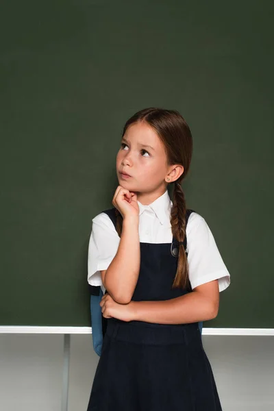 Thoughtful schoolkid looking away and holding hand near face while standing at chalkboard on grey — Stock Photo