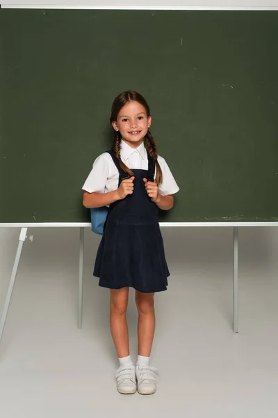Visão de comprimento total de escolar com mochila azul sorrindo para a câmera perto de quadro-negro em cinza — Fotografia de Stock