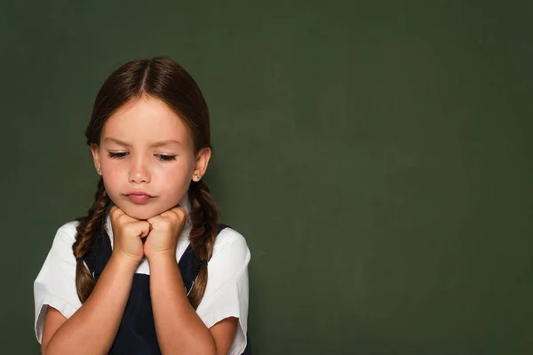 Upset schoolgirl holding hands near chin while standing near chalkboard — Stock Photo