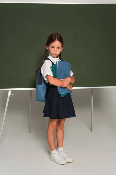 Colegiala ofendido con libros de texto mirando a la cámara cerca de pizarra en gris - foto de stock