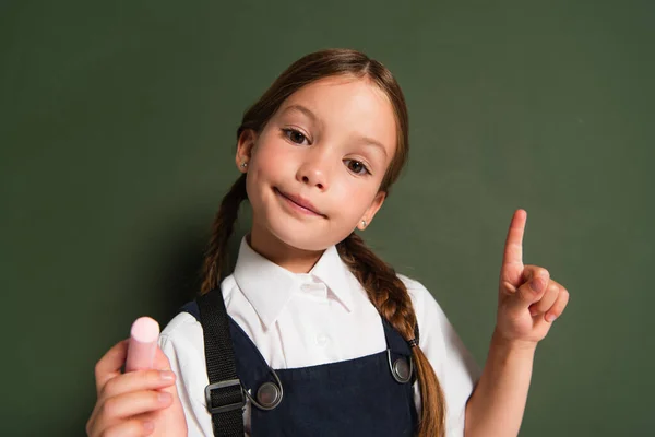 Smiling schoolkid holding chalk and pointing at chalkboard while looking at camera — Stock Photo