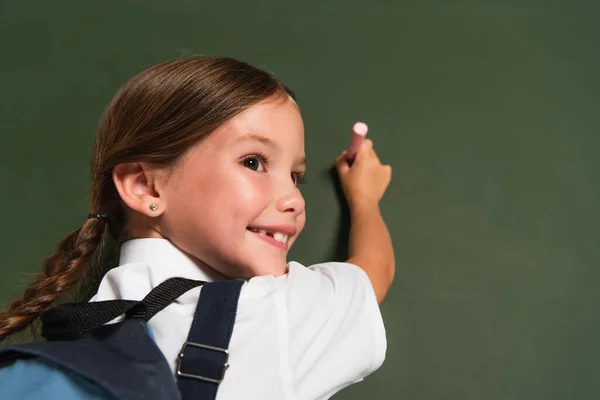 Alegre colegial mirando hacia otro lado mientras escribe en pizarra - foto de stock