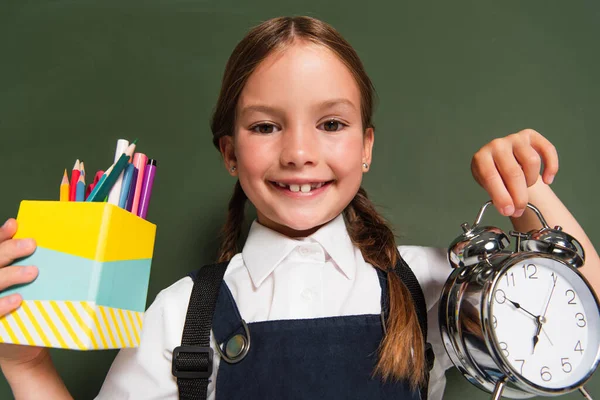 Joyful schoolgirl showing pen holder and alarm clock near chalkboard — Stock Photo