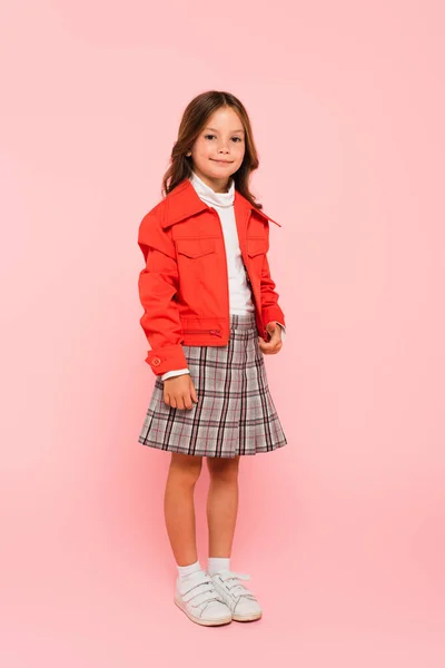 Vista completa de la chica en chaqueta naranja y falda a cuadros mirando a la cámara en rosa - foto de stock