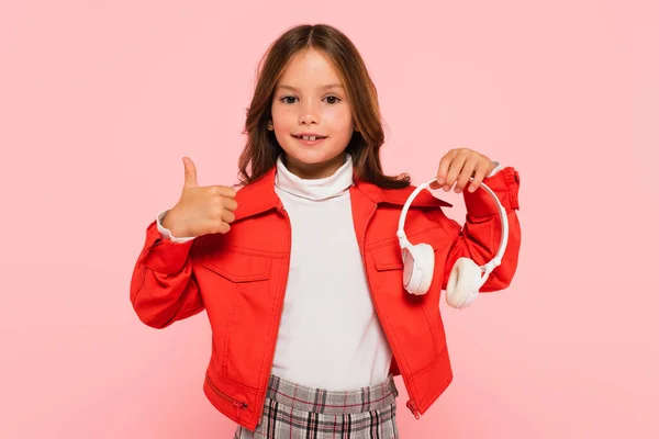 Улыбающаяся девушка в стильной оранжевой куртке держа наушники и показывая большой палец вверх изолированы на розовый — стоковое фото