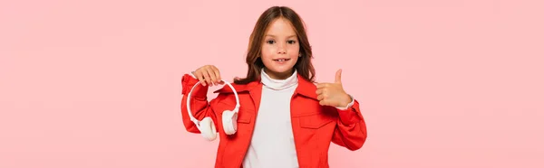 Elegante chica sosteniendo los auriculares y mostrando el pulgar hacia arriba aislado en rosa, bandera - foto de stock