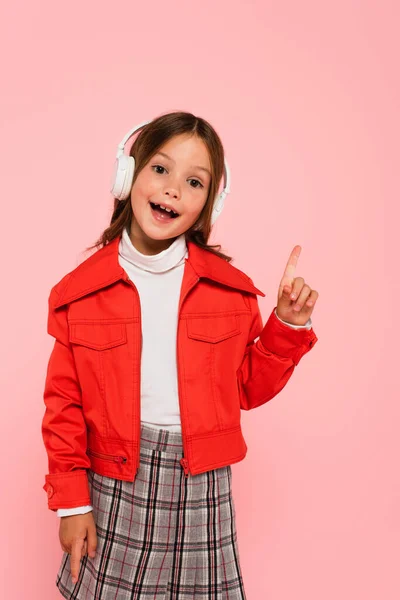 Allegra ragazza in giacca arancione e cuffie puntate con dito isolato su rosa — Foto stock