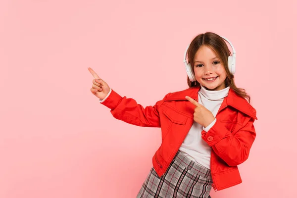 Chica sonriente en auriculares apuntando con los dedos aislados en rosa - foto de stock