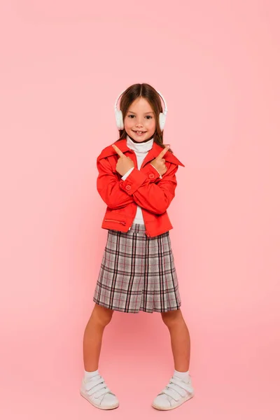 Chica en ropa de moda y auriculares apuntando con los dedos en rosa - foto de stock