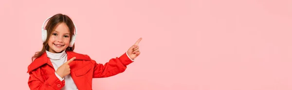 Criança elegante e alegre em fones de ouvido apontando com dedos isolados em rosa, banner — Fotografia de Stock
