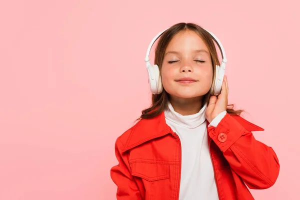 Niño complacido en chaqueta naranja escuchando música con los ojos cerrados aislados en rosa - foto de stock