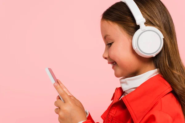 Vista lateral de niño feliz en auriculares con teléfono inteligente aislado en rosa - foto de stock