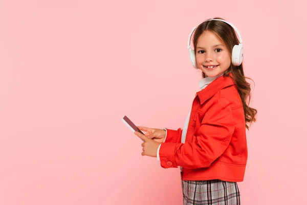 Menina elegante em fones de ouvido usando smartphone isolado em rosa — Fotografia de Stock
