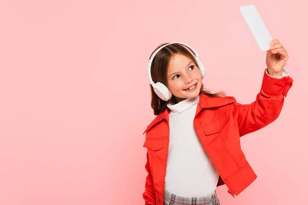 Щаслива і модна дівчина бере селфі в навушниках ізольована на рожевому — стокове фото