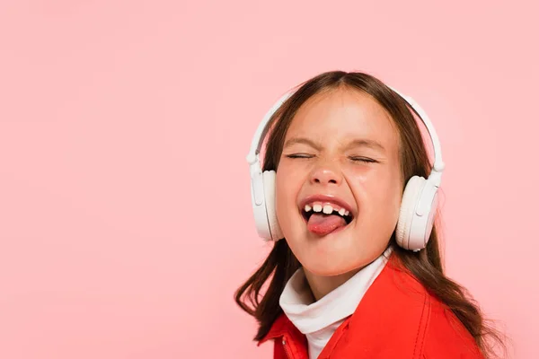 Girl in headphones sticking out tongue with closed eyes isolated on pink — Stock Photo