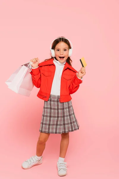 Chica sorprendida en los auriculares de pie con tarjeta de crédito y bolsa de compras en rosa - foto de stock