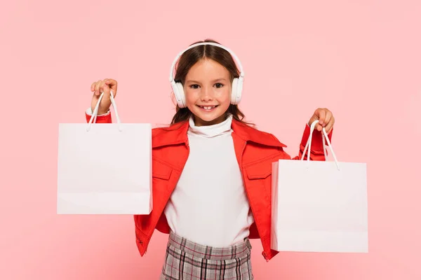 Fröhliches Kind in Kopfhörer und orangefarbener Jacke mit Einkaufstaschen in rosa — Stockfoto