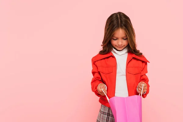 Chica en chaqueta naranja elegante mirando en el bolso aislado en rosa - foto de stock